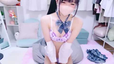 Video of an Asian woman in a plaid bow tie and a floral bra, kneeling on a pink rug in a room with light furniture, wearing a face mask.
