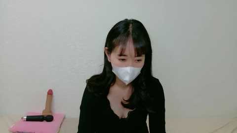 A video of an Asian woman with long black hair, wearing a surgical mask and black top, sitting on a bed. A pink box and dildo are visible on a bedside table.