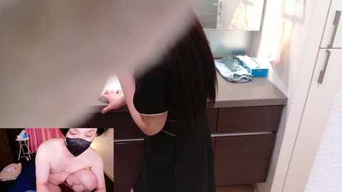 Video of a nude woman with large breasts, wearing a black mask, sitting on a bathroom counter, with a blurred figure in the background.