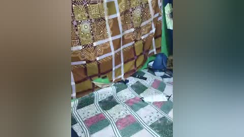 Video of a colorful, patterned blanket with geometric designs and a tapestry wall in the background, featuring intricate mandala-like designs. The blanket is laid out on a green floor, with a blue bag and a pair of shoes partially visible.