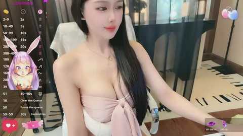A video of an East Asian woman with long black hair and a pale complexion, wearing a pink strapless dress, sitting indoors. The background features a glass door and a white towel.