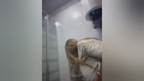 Video of a blonde woman in a beige jacket, looking down, standing in a modern bathroom with white walls and a glass shower door.