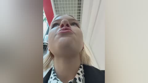 A video of a blonde woman with fair skin, wearing a black-and-white patterned blouse, looking upward with a neutral expression, in a modern indoor setting with white walls and a red object in the background.