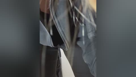 Video of a person in a dark room, wearing a grey jacket and black gloves, with a zipper partially open. Background is out of focus, creating a sense of mystery and suspense.