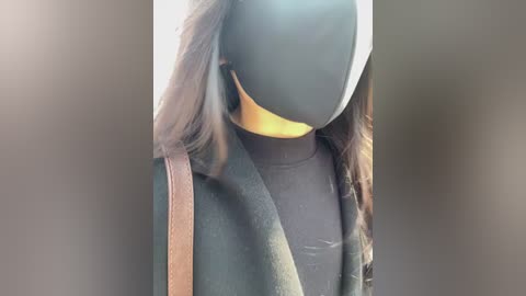 Video of a person with a dark, featureless mask covering the face, wearing a black coat with a fur-lined hood and a brown leather strap over the shoulder, standing in a blurred, neutral-toned background.