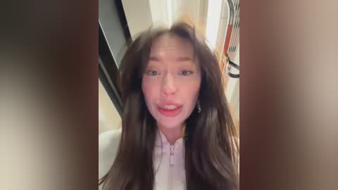 Video of a smiling Asian woman with long, straight brown hair, wearing a white shirt, taken indoors near a door.