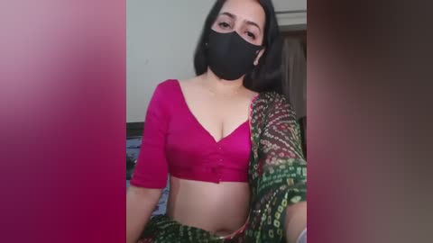Video of a young woman with light skin, dark hair, wearing a black mask, magenta crop top, and a green saree. She stands indoors with a blurred background, suggesting a casual, possibly intimate setting.