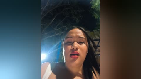 Video of a young Asian woman with long black hair and light skin, wearing a white top, captured outdoors at night with a blurred tree and green foliage in the background.