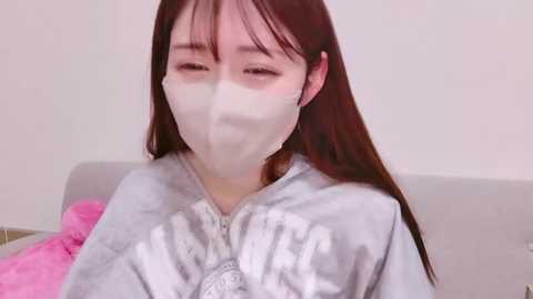 Video of an East Asian woman with long brown hair and pale skin, wearing a white surgical mask and a light blue hospital gown, seated on a light-colored couch.