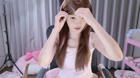 A video of a young Asian woman with long brown hair, sitting in a chair, adjusting her hair with a comb, wearing a pink dress. The background features light gray curtains and pink furniture.