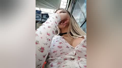 Video of a young woman with light skin and blonde hair, wearing a floral-patterned blouse and a black choker, resting her chin on her hand. Background includes a blurred interior with industrial elements.