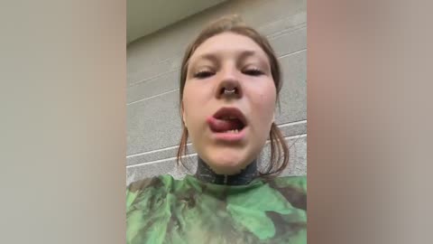 Video of a fair-skinned woman with brown hair, wearing a camouflage-patterned top, sticking her tongue out, set against a concrete wall background.