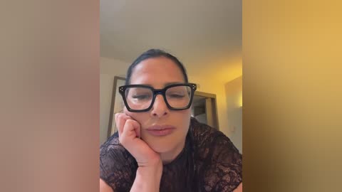 Video of a woman with medium skin tone, dark hair tied back, wearing black rectangular glasses and a black lace top, resting her chin on her hand, indoors with a beige wall and a door in the background.