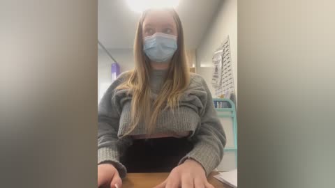 Video of a young woman with light skin, long blonde hair, and a blue surgical mask, wearing a gray sweater, sitting at a wooden desk in a classroom.