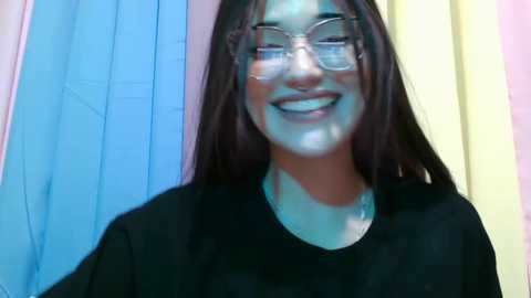 A young woman with long, dark hair, wearing glasses and a black shirt, grins widely with white powder on her face, set against a pastel-colored backdrop.