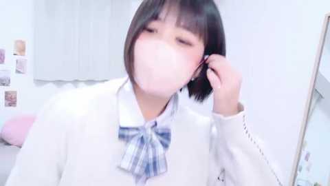 A young Asian woman with short black hair and a white mask, wearing a white sweater and blue plaid bow tie, adjusts her earpiece in a minimalist, brightly lit room with white walls and a pink pillow.