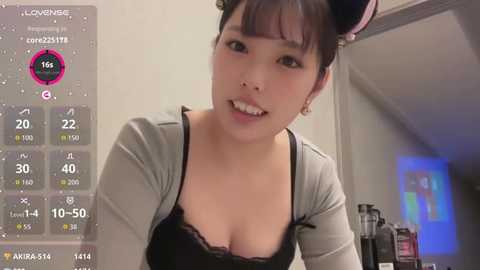 Video of an East Asian woman with short black hair in a bun, wearing a black lace bra and gray cardigan, smiling in a cozy indoor setting with a bathroom mirror in the background.