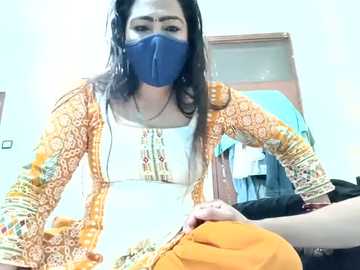 Video of a woman with long dark hair, wearing a blue face mask, orange and white patterned blouse, and a yellow skirt, sitting on a bed in a brightly lit room.