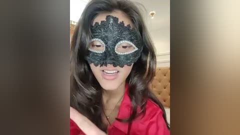 Video of a smiling woman with long dark hair, wearing a black lace masquerade mask with silver embellishments, a red dress, and a beige tufted headboard in the background.