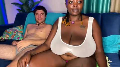 Video of a dark-skinned woman with large breasts and a necklace, sitting on a blue couch with a pale-skinned man behind her, both wearing minimal clothing.