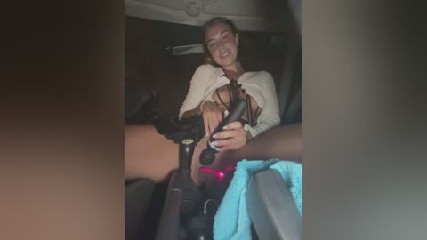 Video of a blonde woman with fair skin, wearing a white top, smiling, holding a black handgun inside a car.