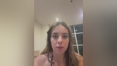 Video of a young Caucasian woman with long, wavy brown hair, light skin, and a neutral expression, wearing a purple necklace, indoors, with a white window and beige walls in the background.