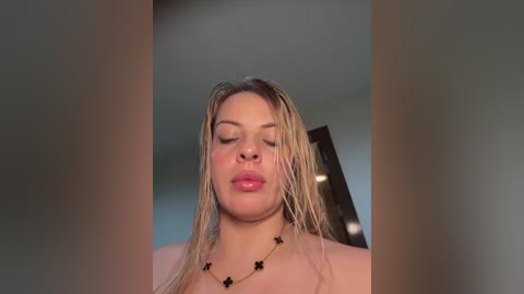A video of a nude, fair-skinned woman with wet blonde hair, wearing a black star necklace, standing indoors, eyes closed, lips slightly parted, and a serene expression.