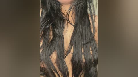 A close-up video of a woman's chest with long, wavy black hair cascading over her bare breasts. The background is a blurred, neutral beige tone, emphasizing the dark hair and skin tones.
