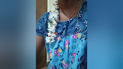 Video of a woman's torso wearing a vibrant blue floral blouse with a white floral necklace, standing in a beige room.