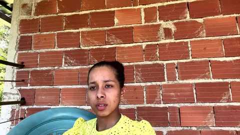 Video of a young woman with medium brown skin and dark hair tied back, wearing a yellow knitted sweater, seated against a red brick wall with a blue cushion visible on her left.