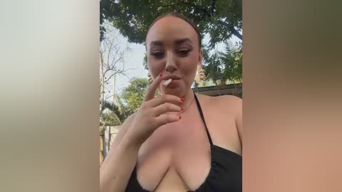 A video of a bald, light-skinned woman with a slender physique, wearing a black halter top, posing provocatively outdoors with a blurred background of greenery and trees.