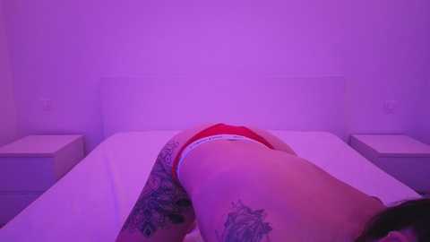 A video of a person with light skin and tattoos, lying face down on a bed in a dimly lit room, wearing red underwear. The background is purple-hued.