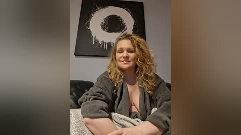Video of a middle-aged, plus-size, curly-haired woman in a grey robe, sitting on a couch. Behind her, a black abstract painting with a white circle is mounted on a grey wall.