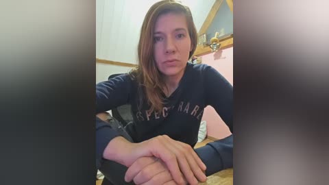 Video of a young woman with light skin and brown hair, wearing a dark blue \"Super Mario\" sweatshirt, sitting on a bed, indoors with a blurred background.