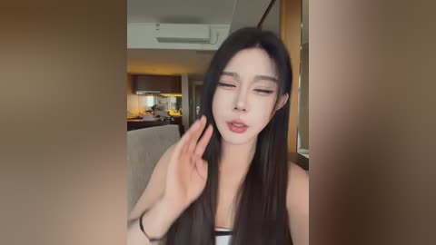 Media: A video of an East Asian woman with long black hair, wearing a striped top, standing indoors, touching her face, in a modern kitchen with wooden cabinets and beige walls.