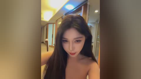 Video of an Asian woman with long black hair, fair skin, and red lipstick, partially nude, standing in a modern, dimly lit hallway with wooden doors and warm lighting.