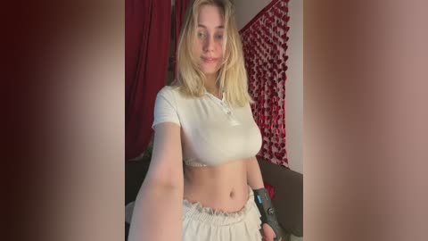 Video of a blonde woman in a tight, white crop top, showing her midriff and large breasts, wearing a white skirt and black wristband, standing in a room with red curtains and a patterned wall.