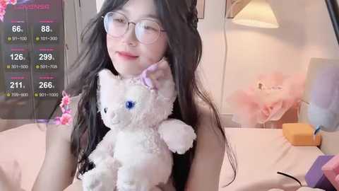 A video of an East Asian woman with long black hair, glasses, and a white teddy bear, sitting on a bed with pink sheets and a floral backdrop.