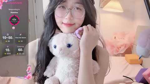 Video of an Asian woman with long black hair, glasses, and a white teddy bear, smiling in a soft-lit room with pink floral decor and a lamp.