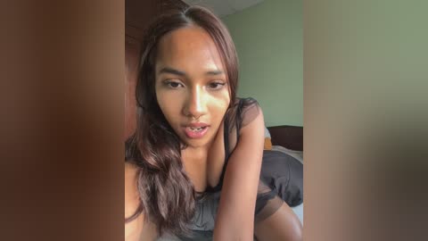 Video of a young, light-skinned woman with long dark hair, wearing a black tank top, lying on a bed with a green wall behind her. She has a subtle, pouty expression.