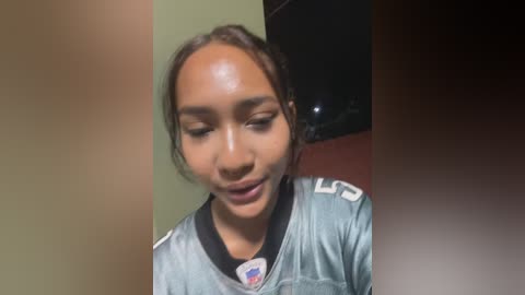 Video of a young Black woman with a light brown complexion, wearing a silver and black football jersey, smiling. Background features a green wall and a dark object.