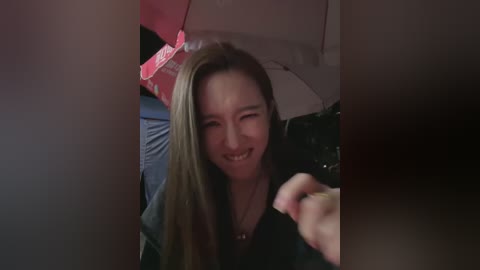 A candid video of a young woman with long, straight brown hair and a joyful smile, captured in a dimly lit room with a red and white patterned bedspread and a black camera in the foreground.