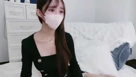 Video of an East Asian woman with long brown hair, wearing a white mask, black long-sleeve top, and sitting on a white bed in a white room with drawers and a gray pillow.