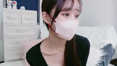 Video of a young Asian woman with long black hair, wearing a white surgical mask, black top, and gold necklace, indoors, near white dresser with toys, white bed, and fluffy pillows.