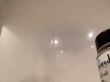 Video of a glossy white ceramic tile backsplash with a vertical seam, reflecting soft light from recessed ceiling lights. Partial view of a black and white \"Lipton\" tea container on the right.