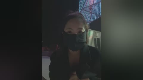 Video of a person with a black mask, wearing a black jacket, and standing in a dimly lit room with a colorful, geometric-patterned ceiling light.