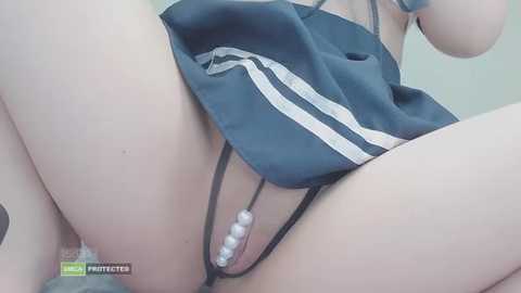 Video of a light-skinned woman in black lingerie, revealing her genitals adorned with pearl beads. She has a slim physique and wears a sports bra. The background is a plain, light-colored wall.