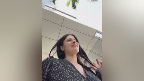 Media: A video of a young woman with long dark hair, wearing a black polka-dotted dress, standing under a modern building with a white ceiling and palm trees in the background.