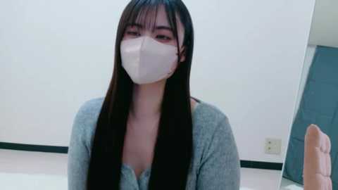 Video of an East Asian woman with long black hair and fair skin, wearing a white face mask and a light gray sweater, indoors with white walls and a blue door in the background.