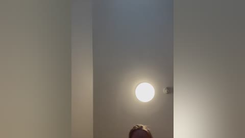 Video of a dimly lit room with a round, bright light shining from a door in the center, casting a soft glow on a person's head, whose face is not visible. The walls are plain and white.